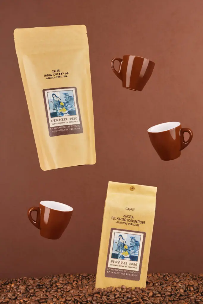 Confezioni di caffè artigianale Penazzi 1926 con tazzine sospese e chicchi sul fondo, simbolo della passione per la tostatura tradizionale