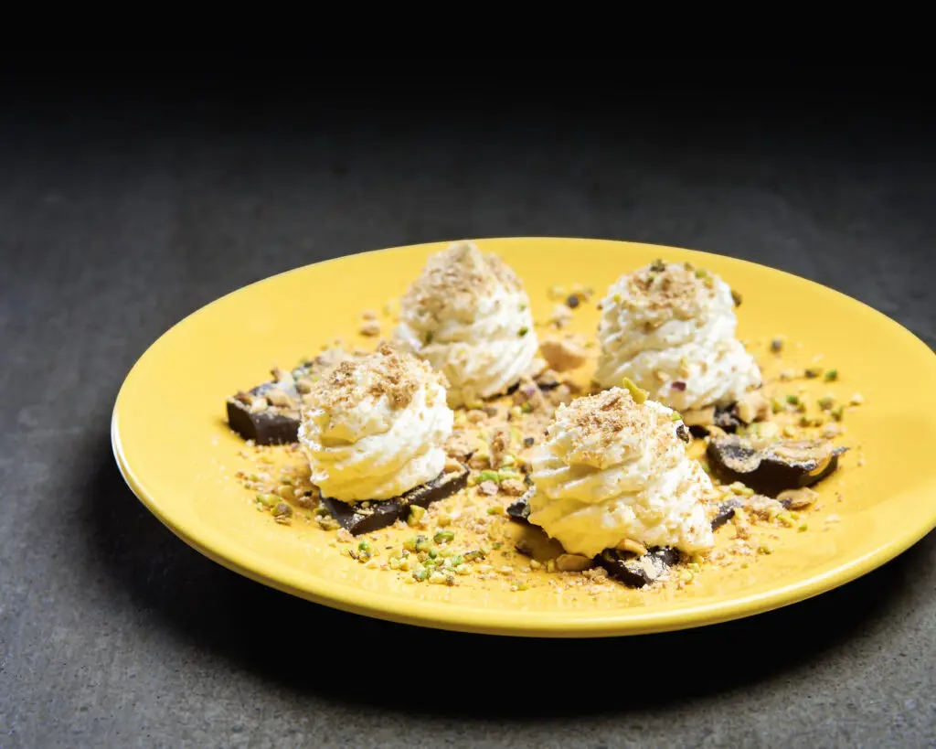 Dessert su piatto giallo con ciuffi di crema e granella di pistacchio su fondo scuro
