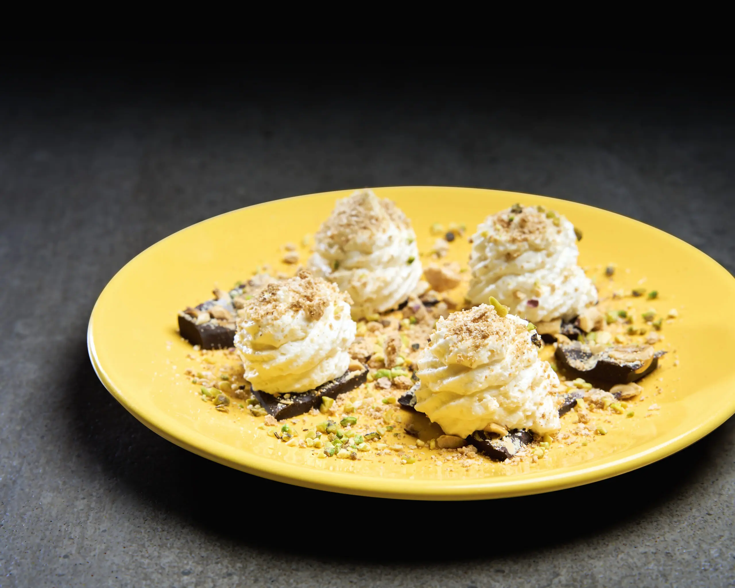 Dessert su piatto giallo con ciuffi di crema e granella di pistacchio su fondo scuro
