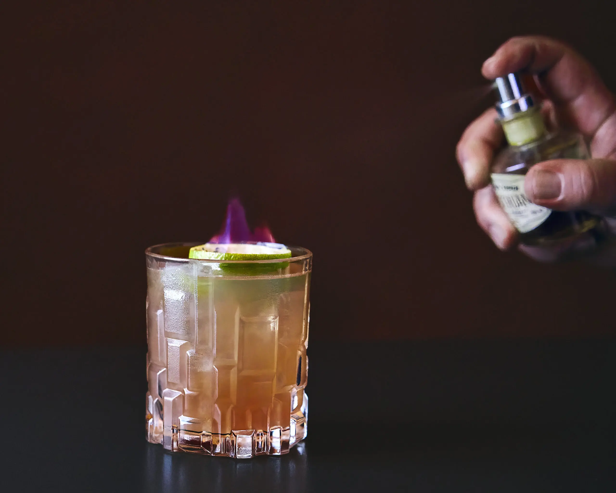 Mano che spruzza aroma su cocktail in bicchiere basso con fiamma su sfondo scuro